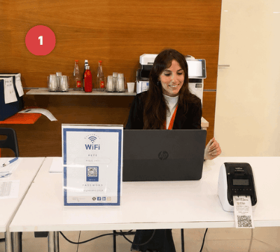 Persona en una mesa de acreditación con laptop, impresora y código QR para registrar participantes.